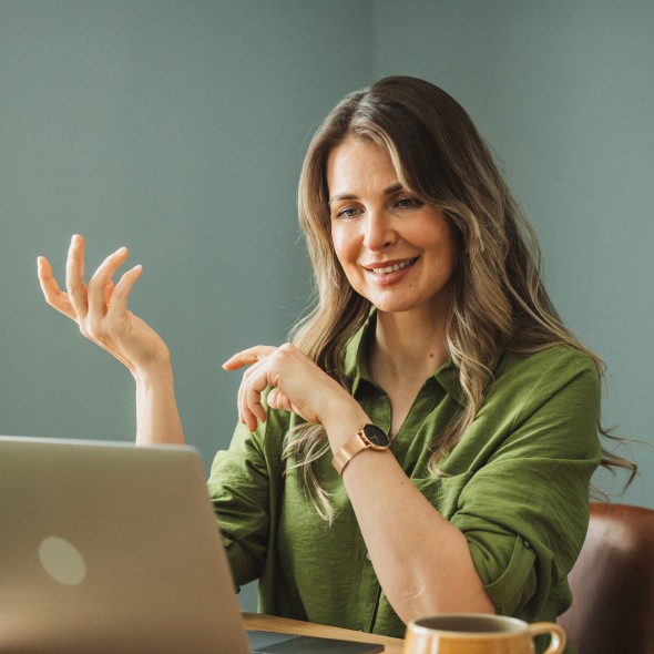 A woman seated at a table, focused on her laptop while engaged in conversation.