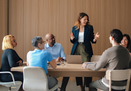 Group of people participating in a meeting around a conference table while one person presents to the group in a modern boardroom.