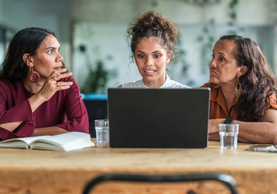 Three people gathered around a laptop at a table, collaborating in an office setting.