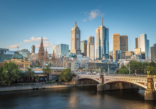 A river flows beneath a bridge, with tall buildings rising in the background against a clear sky in Melbourne.