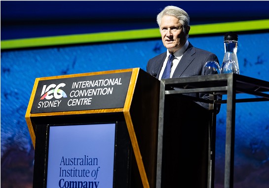 A man in a suit and tie stands confidently at a podium, speaking to an audience.