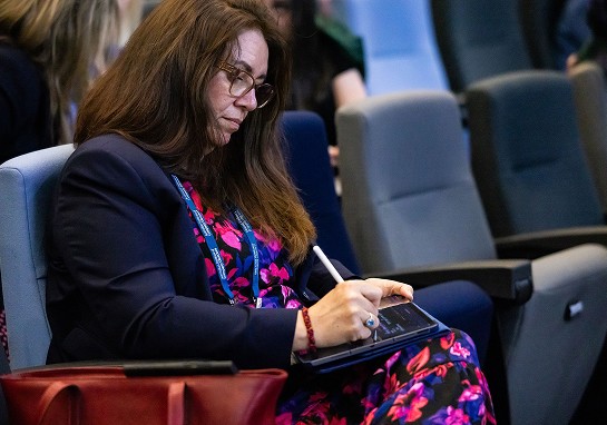 A woman in a vibrant floral dress sits comfortably in a chair, taking notes at an event.