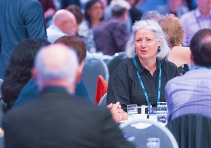 A woman at a table, engaged in conversation, with others visible in the background.