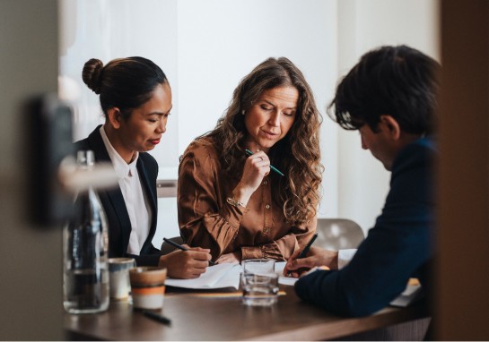 A group of three business individuals at a table, engaged in conversation while examining papers.
