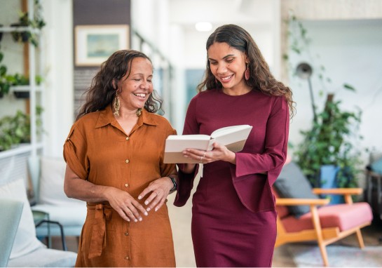 Two smiling women stand in a room, one holding a book while the other listens attentively.