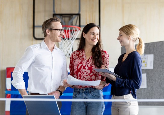 Three people engaged in conversation while standing infront of a basketball ring.