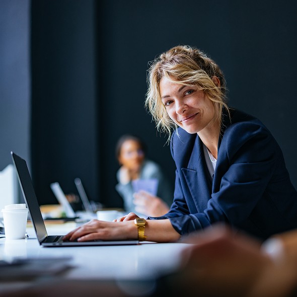 A woman in a business suit smiles while working on her laptop in a professional setting.
