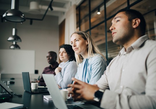 A group of diverse individuals seated at a table, each using a laptop, engaged in discussion or collaboration.