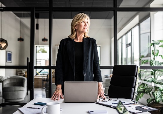 A woman stands at a desk with her laptop, in a professional workspace setting.