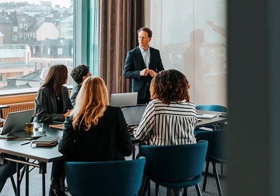 A man dressed in a suit stands before a group, presenting information and engaging with his audience.