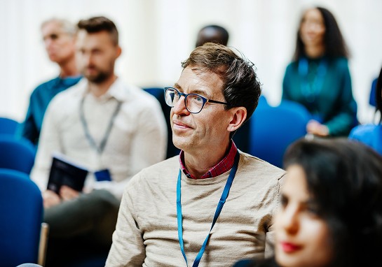 A man wearing glasses sits with a group of people, engaged in discussion or presentation.