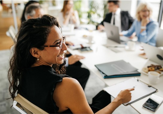A woman with a bright smile sits at a table with several others in an office setting.